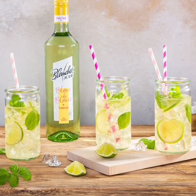 Drei Gläser mit erfrischenden Cocktails, garniert mit Limetten und frischem Basilikum, stehen auf einem Holztisch.