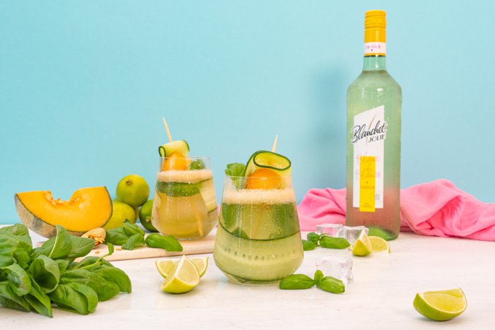 Zwei erfrischende Cocktails mit Melone, Limette und Gurke stehen auf einem Tisch, umgeben von frischem Basilikum und Limettenscheiben.