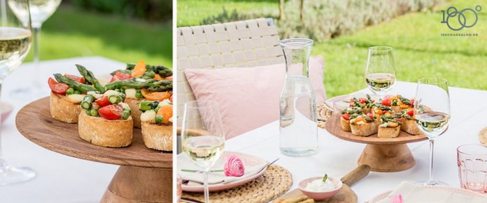 Ein rustikales Holztablett mit bunten Gemüsetoasts steht auf einem eleganten Tisch im Freien, umgeben von Gläsern und einer Karaffe.