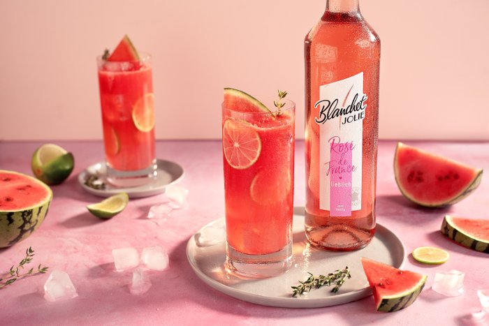 Zwei Gläser mit einem erfrischenden Wassermelonen-Drink stehen auf einem rosa Tisch, umgeben von frischen Früchten und Eiswürfeln.