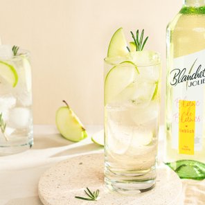 Ein erfrischendes Getränk mit Apfelscheiben und Rosmarin, serviert in einem hohen Glas auf einem hellen Tisch.