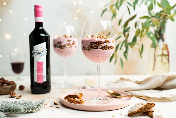 Zwei elegante Gläser mit rosa Dessert, geschichtet mit Keksen, stehen auf einem Tisch neben einer Flasche Wein und Nüssen.