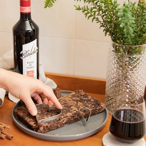 Eine Hand schneidet ein Stück Schokoladenkuchen mit einem herzförmigen Ausstecher auf einem grauen Teller.