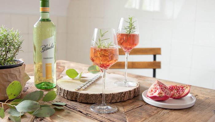 Zwei Gläser mit rosafarbenen Getränken, dekoriert mit Rosmarin, stehen auf einem rustikalen Holztisch neben einer Flasche Chardonnay von Blanchet.