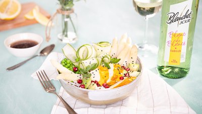 Eine kreative Salatkomposition mit frischen Zutaten, darunter Spargel und Gurken, neben einer Flasche Blanc de Blancs Wein.