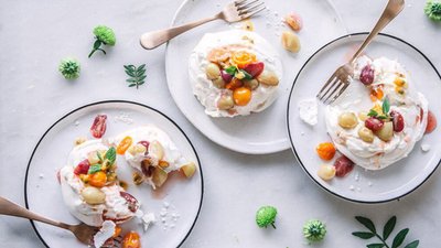Drei Teller mit Pavlova, garniert mit bunten Früchten wie Trauben und Kirschen, umgeben von frischen Kräutern.