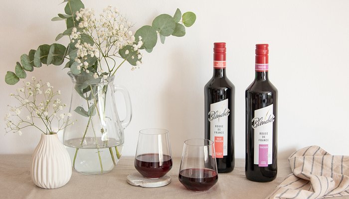 Zwei Flaschen Rotwein von Blanchet stehen neben einem Krug mit frischen Blumen und einem Glas Wein auf einem hellen Tisch.