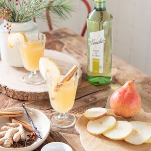 Zwei Gläser mit einem fruchtigen Getränk, garniert mit Apfelscheiben und Zimtstangen, stehen auf einem rustikalen Holztisch.