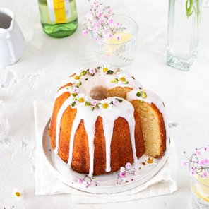 Ein saftiger Gugelhupf mit einer glänzenden Zuckerglasur, dekoriert mit kleinen Blumen und auf einem weißen Teller serviert.
