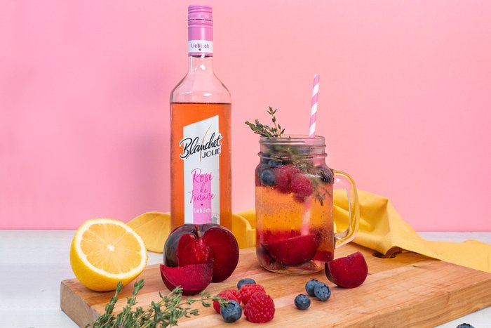 Ein Glas mit einem erfrischenden Getränk, gefüllt mit Beeren und einem Strohhalm, steht vor einer rosa Wand.