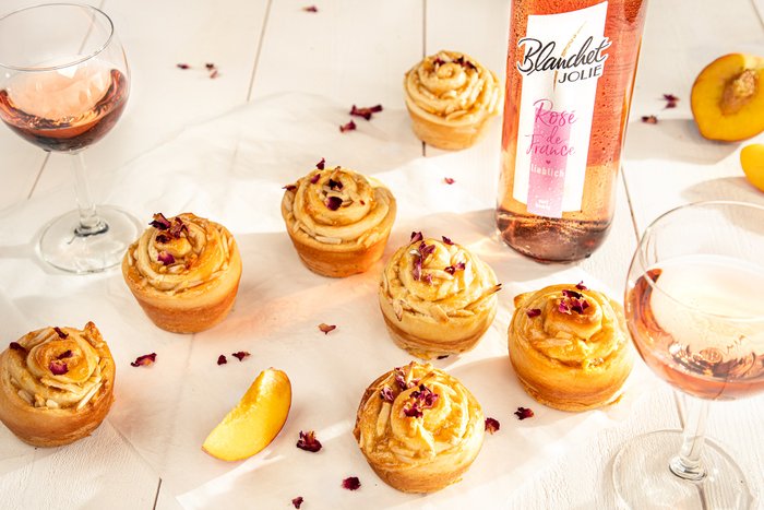 Sechs kunstvoll dekorierte Cupcakes mit Rosenblättern stehen auf einem weißen Tisch, umgeben von Gläsern mit Roséwein.