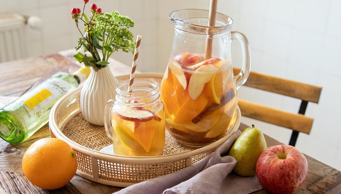 Ein rustikaler Tisch mit einer Karaffe gefüllt mit fruchtigem Getränk, umgeben von frischen Früchten und einer kleinen Vase.