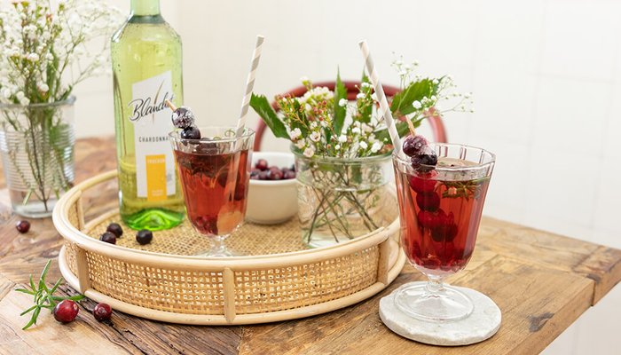 Zwei Gläser mit einem roten Getränk, dekoriert mit Beeren, stehen auf einem Tablett aus Rattan neben einer Flasche Wein von Blanchet.