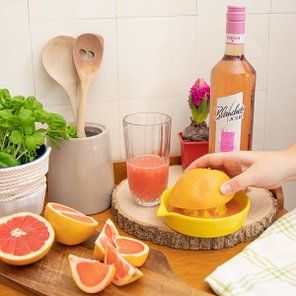 Eine Hand presst eine Grapefruit in eine gelbe Saftpresse, während frische Zutaten und ein Glas Saft im Hintergrund stehen.