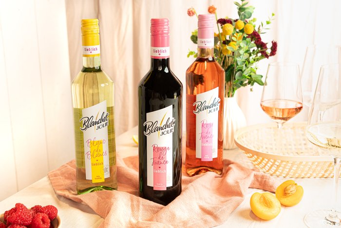 Auf einem Tisch liegen drei Flaschen des Weins 'Blanchet Jolie' in Gelb, Schwarz und Rosé, neben frischen Himbeeren und Aprikosen.