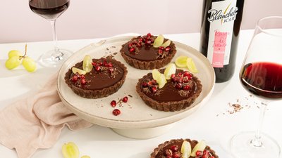 Drei köstliche Schokoladentörtchen mit Granatapfelkernen und Trauben auf einem hellen Teller, umgeben von einem weichen Tuch.