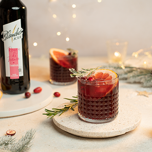 Zwei Gläser mit einem dunklen Getränk, garniert mit Grapefruit und Beeren, stehen auf einem hellen Tisch mit Lichtern im Hintergrund.