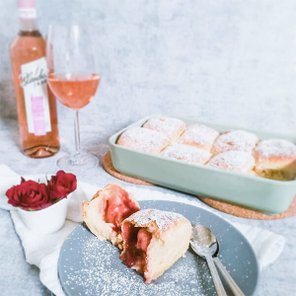 Ein Teller mit einem Stück fruchtigem Dessert, bestäubt mit Puderzucker, neben einer Flasche Roséwein und einer Weinglas.