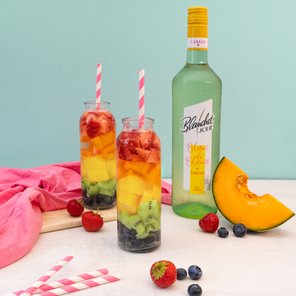 Zwei Gläser mit buntem Obstsalat, gefüllt mit Erdbeeren, Kiwi und Melone, stehen auf einem Tisch neben einer Flasche Blanchet Wein.