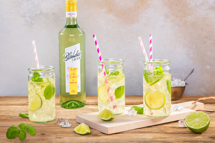 Drei Gläser mit erfrischenden Cocktails, garniert mit Limetten und frischem Basilikum, stehen auf einem Holztisch.