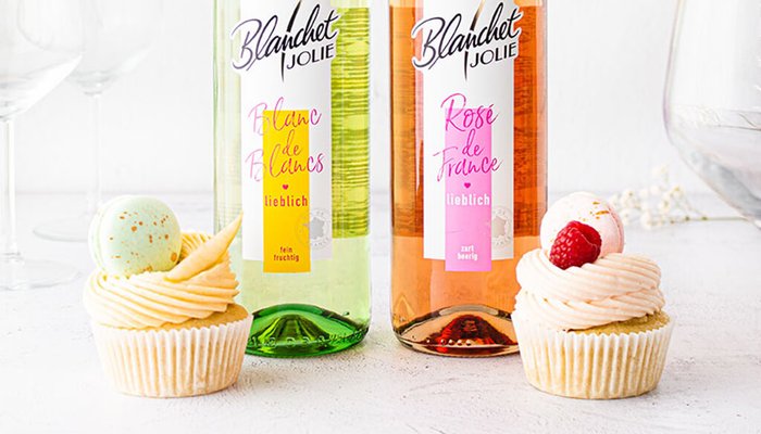 Zwei köstliche Cupcakes mit Sahnehaube stehen neben Flaschen von Blanc de Blancs und Rosé de France auf einem hellen Tisch.