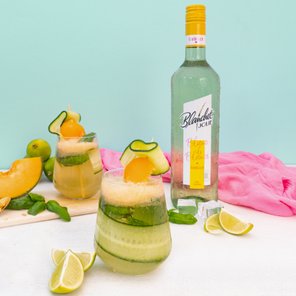 Zwei stilvolle Cocktails mit Melonenstücken und Gurkenscheiben stehen auf einem Tisch, umgeben von Limetten und Eiswürfeln.