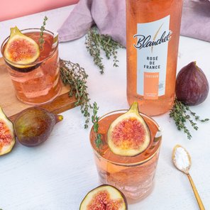 Zwei Gläser mit Roséwein, dekoriert mit frischen Feigen und Thymian, auf einem hellen Tisch arrangiert.