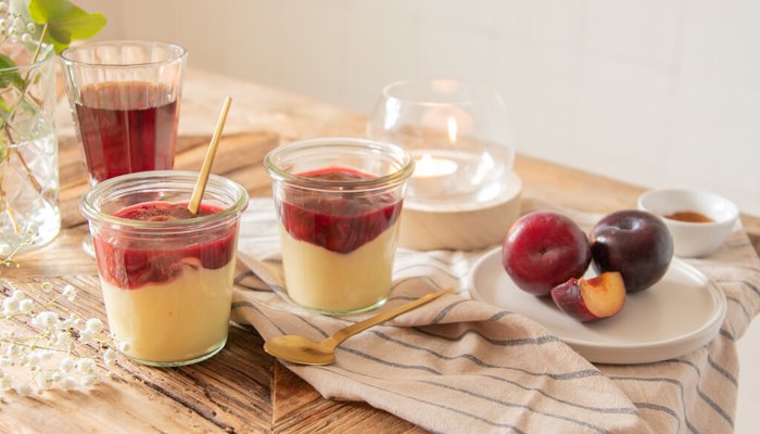 Zwei Gläser mit Pudding, einer mit roter Fruchtschicht, stehen auf einem Holztisch neben frischen Pflaumen und einem Getränk.