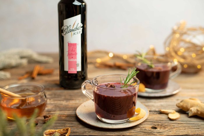 Ein Glas mit einem dunklen Getränk, garniert mit Rosmarin und Zitrusfrüchten, steht auf einem rustikalen Holztisch.