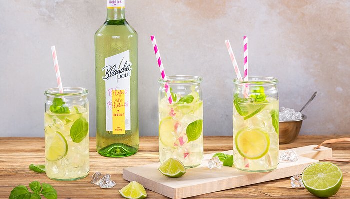 Ein rustikaler Holztisch zeigt drei Gläser mit einem spritzigen Getränk und einer Flasche Wein von Blanchet im Hintergrund.