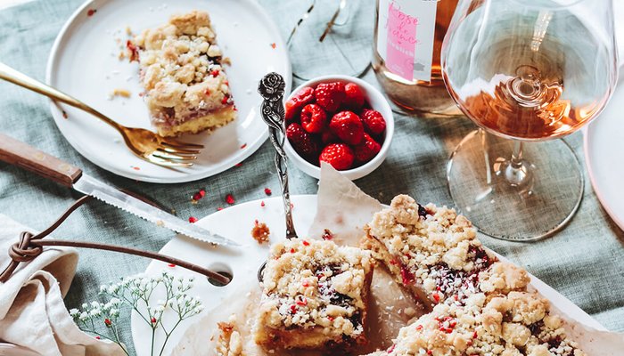Ein köstlicher Erdbeerkuchen mit Streuseln auf einem Teller, umgeben von frischen Himbeeren und einem Glas Roséwein.