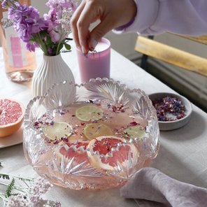 Eine Hand streut essbare Blüten in eine dekorative Schüssel mit fruchtigem Wasser, umgeben von frischen Blumen und Obst.