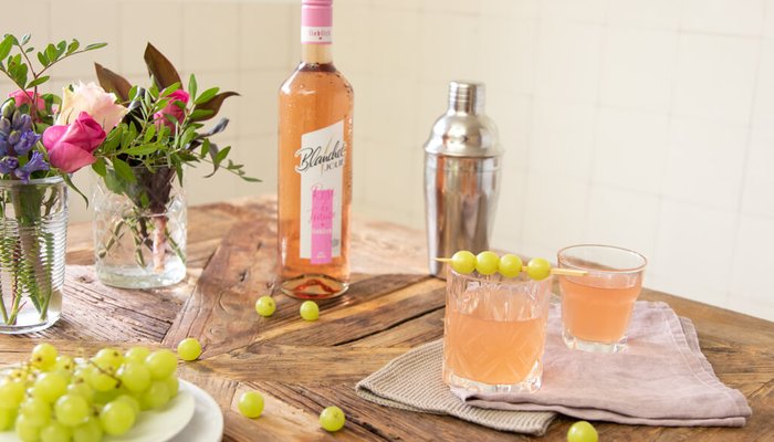 Ein rustikaler Holztisch ist mit einem Glas Roséwein, Cocktails und frischen Trauben dekoriert, umgeben von bunten Blumen.