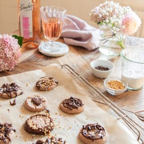 Auf einem Holztisch liegen frisch gebackene Kekse mit Schokoladenglasur und Nüssen, umgeben von Blumen und einem Glas Rosé.