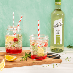 Zwei Gläser mit bunten Strohhalmen und sprudelndem Getränk, umgeben von frischen Kräutern und einer Flasche Wein von Blanchet im Hintergrund.