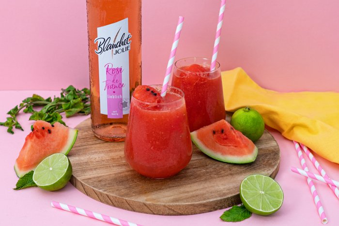 Ein erfrischender Wassermelonen-Smoothie in einem Glas mit einem pinken Strohhalm, neben einer Flasche Rosé von Blanchet und Limetten.