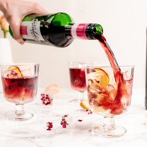 Ein Handgelenk gießt roten Wein in ein Glas mit Eis, umgeben von frischen Blütenblättern und einer Flasche im Hintergrund.